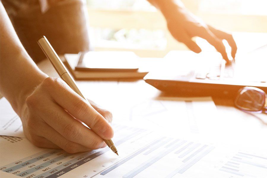 Hands writing on paperwork at a sunlit desk with a laptop and glasses nearby