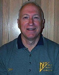 Smiling man in a dark green polo shirt with a yellow logo, standing in front of a wooden door.