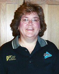 Smiling person in glasses wearing a black polo shirt with logos, standing against a wood-paneled wall