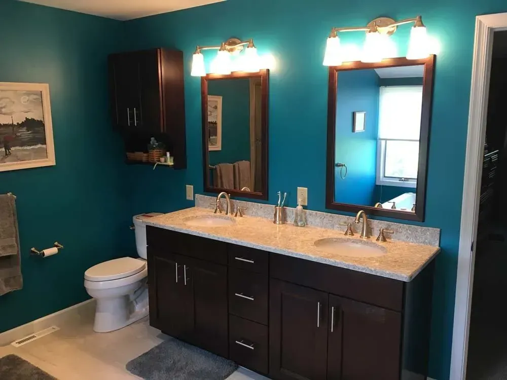 Teal bathroom with double vanity, mirrors, lights, toilet, and window.