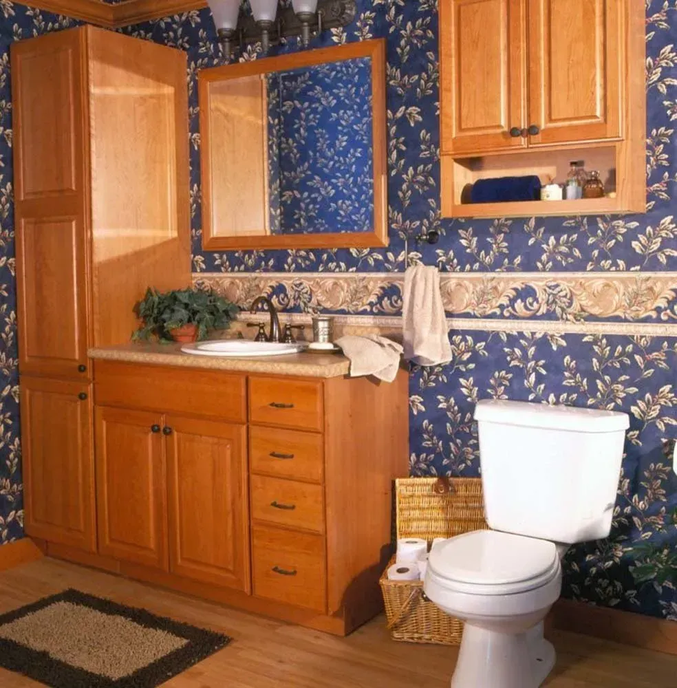 Wood-paneled bathroom with blue floral wallpaper, wooden vanity, toilet, mirror, and rug.