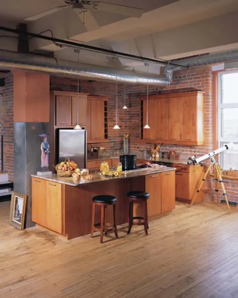 Loft kitchen with wood cabinets, island, stools, exposed brick, and large windows.