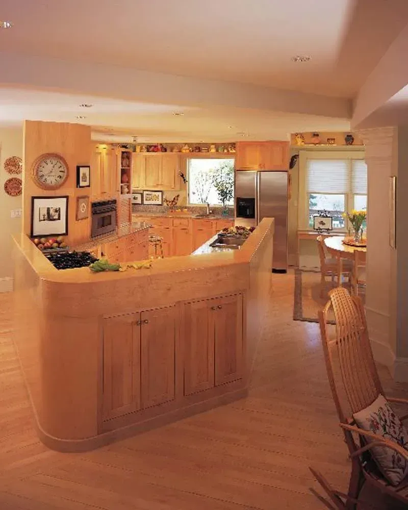 Warm kitchen with light wood cabinets, curved island, stainless fridge, and sunlit dining nook.