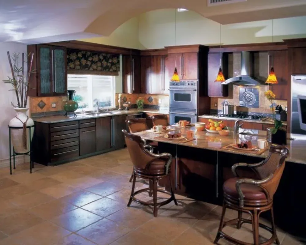 Warm kitchen with dark wood cabinets, island table set for a meal, tiled floor, and pendant lights.
