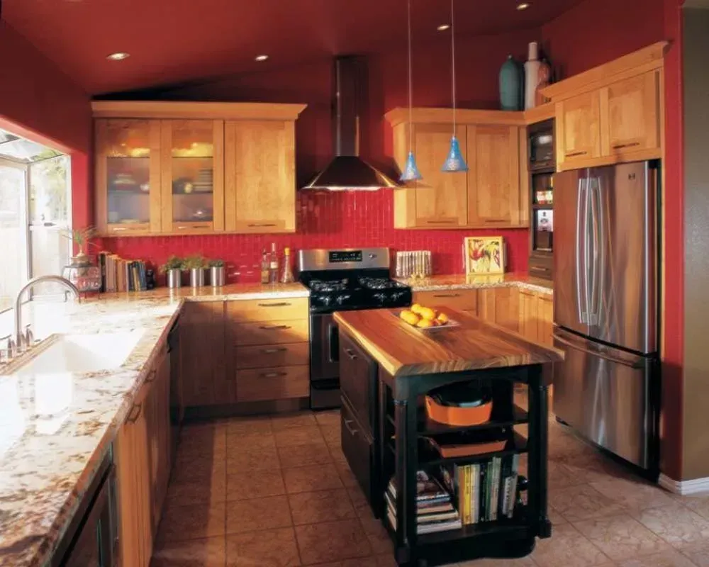 Warm kitchen with wood cabinets, red walls, stainless fridge, and a central island beneath a window.
