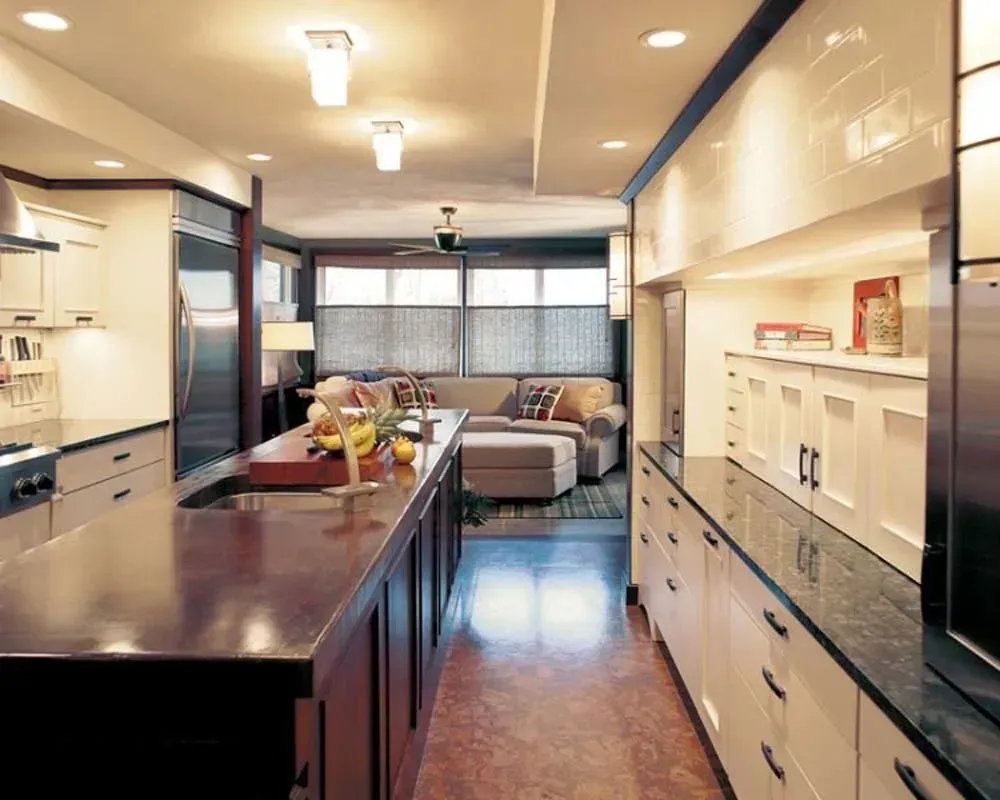 Modern kitchen with a long island, white cabinets, recessed lights, and a bright living area beyond.
