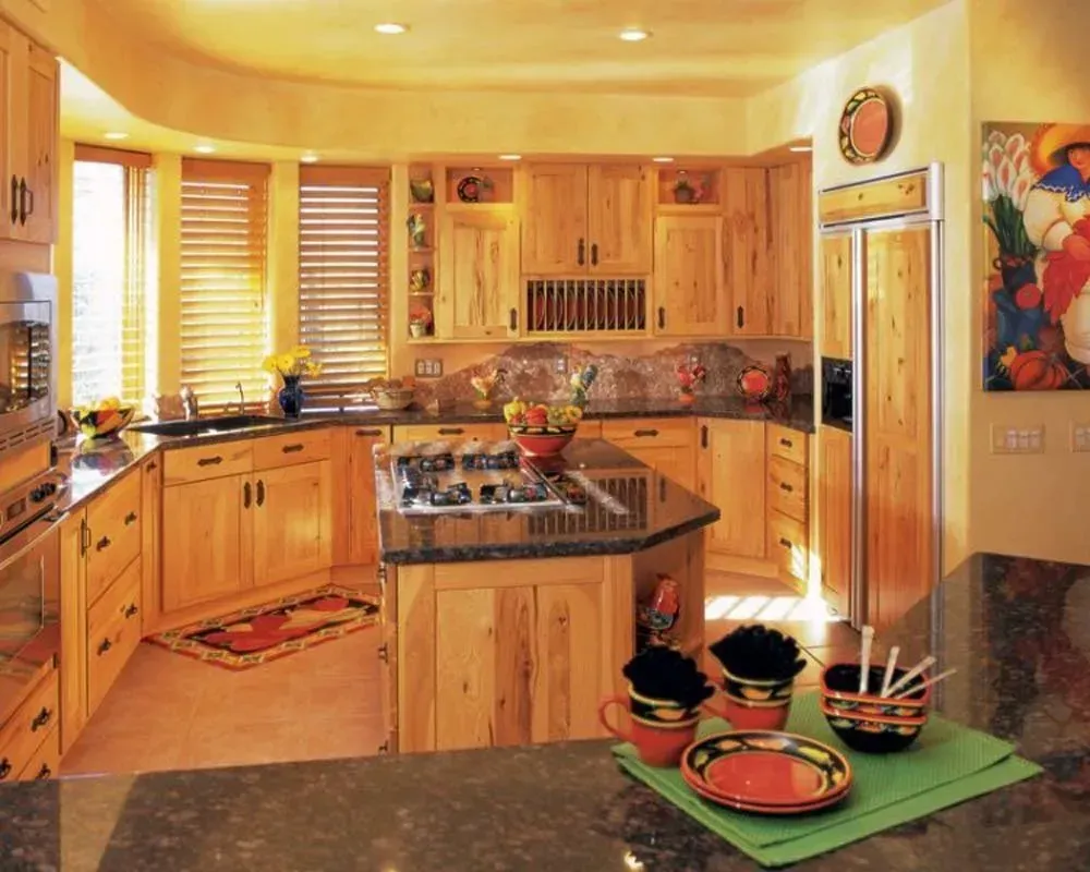 Warm wooden kitchen with island, granite counters, sunlight through blinds, and colorful bowls on a counter.