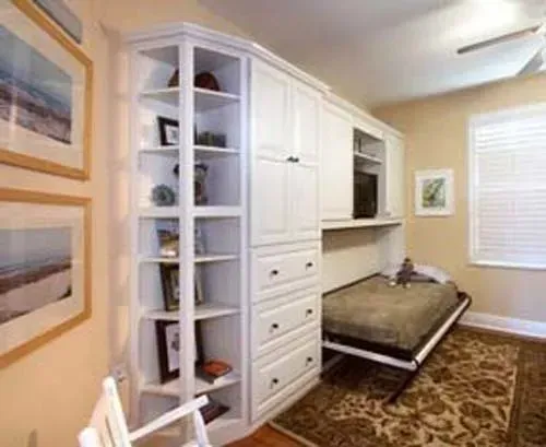 Built-in white shelving and desk in a beige room with framed art, a window, and a patterned rug