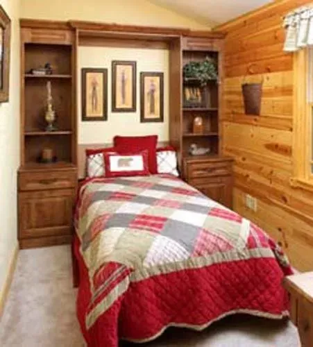 Cozy small bedroom with wood-paneled walls, single bed in red and gray quilt, and built-in shelves and cabinets