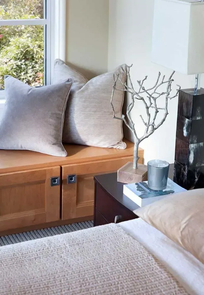 Cozy bedroom corner with window seat, gray pillows, beige bed, and a nightstand with decor