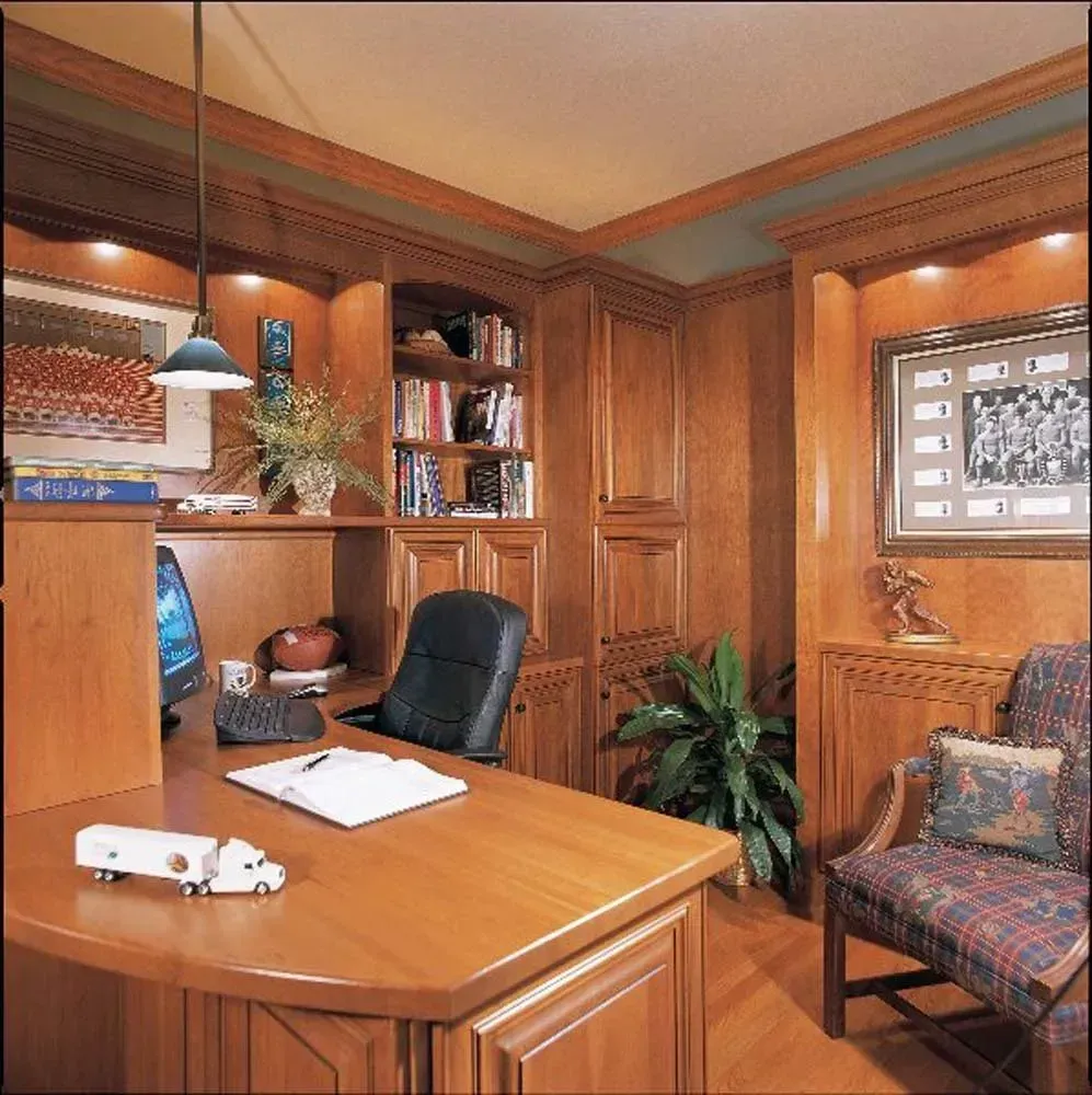 Wood-paneled home office with a desk, bookshelves, black chair, and patterned armchair near a window.