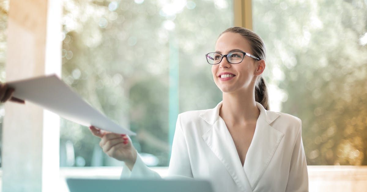 Smiling woman in glasses holding papers in a bright office window.