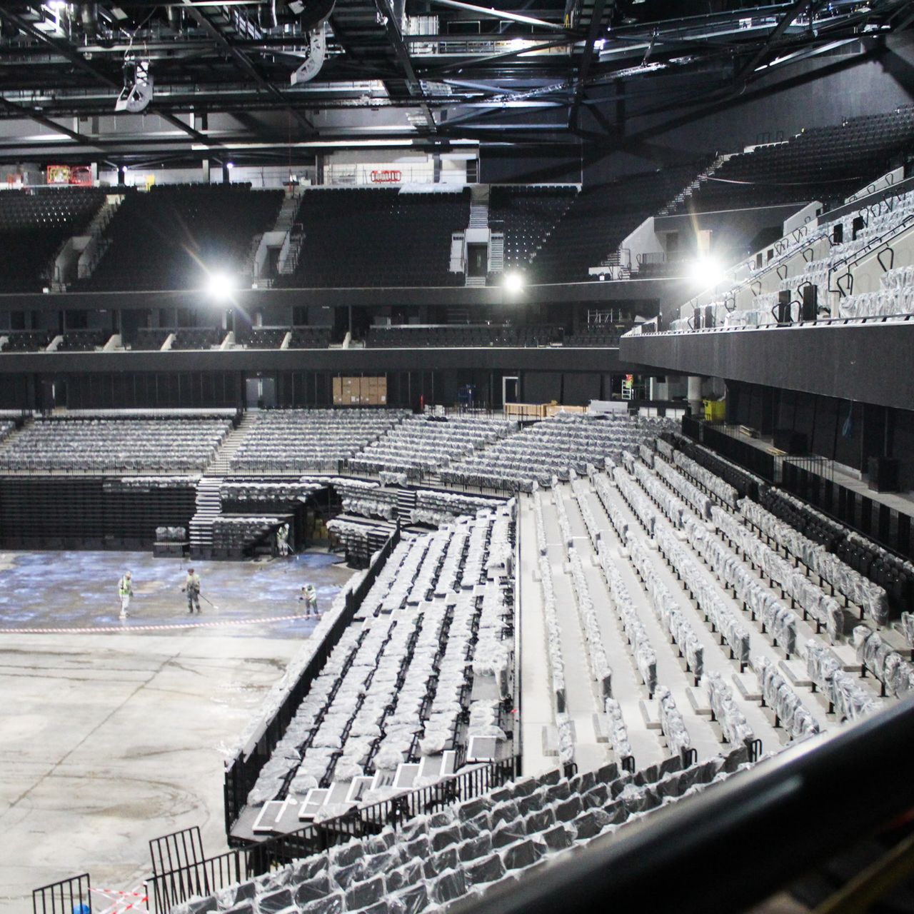 Empty arena under construction, gray seating, workers on the floor, overhead lighting.