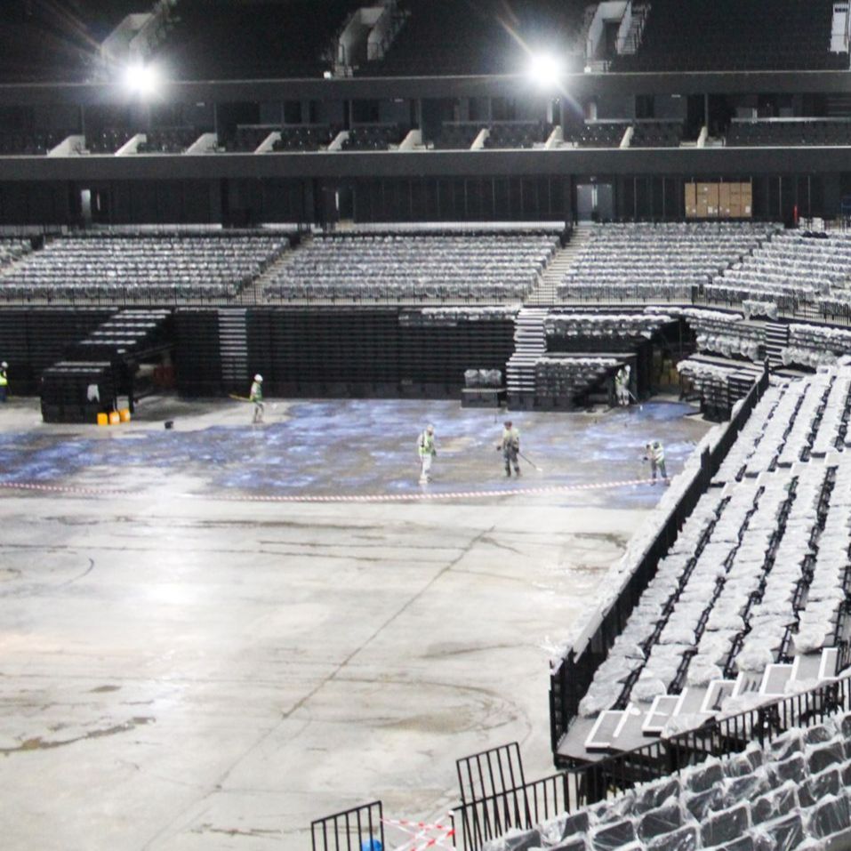 Workers cleaning a large concrete arena floor, surrounded by seating in preparation for an event.