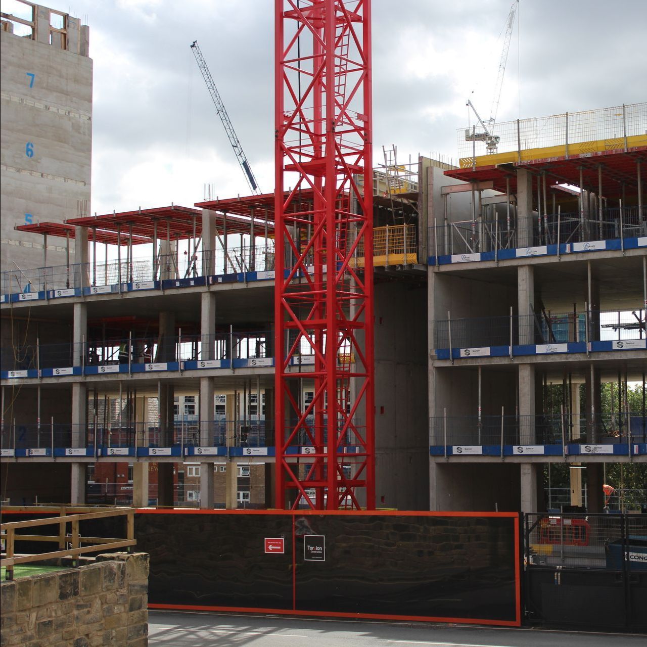 Construction site with red crane; building frames, scaffolding, and fencing. Cloudy sky.