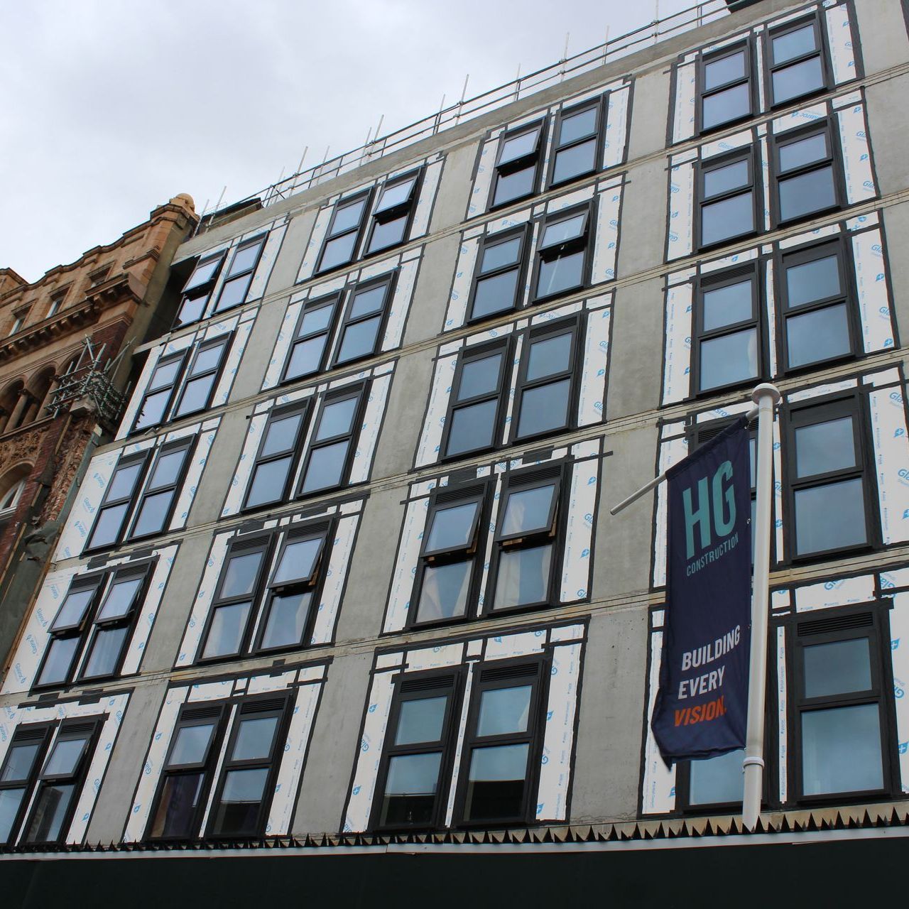 Apartment building under construction with multiple windows, a blue and white HG banner, and a cloudy sky.