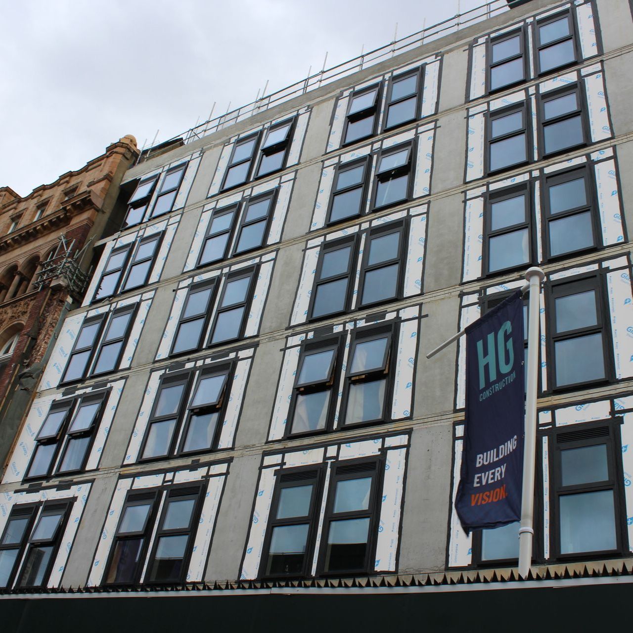 Gray multi-story building with many rectangular windows, construction work visible. HG flag in the foreground.