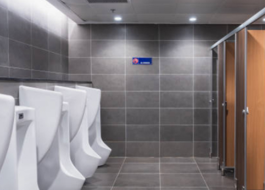 Public restroom with urinals on the left and stalls on the right, tiled walls, neutral tones.