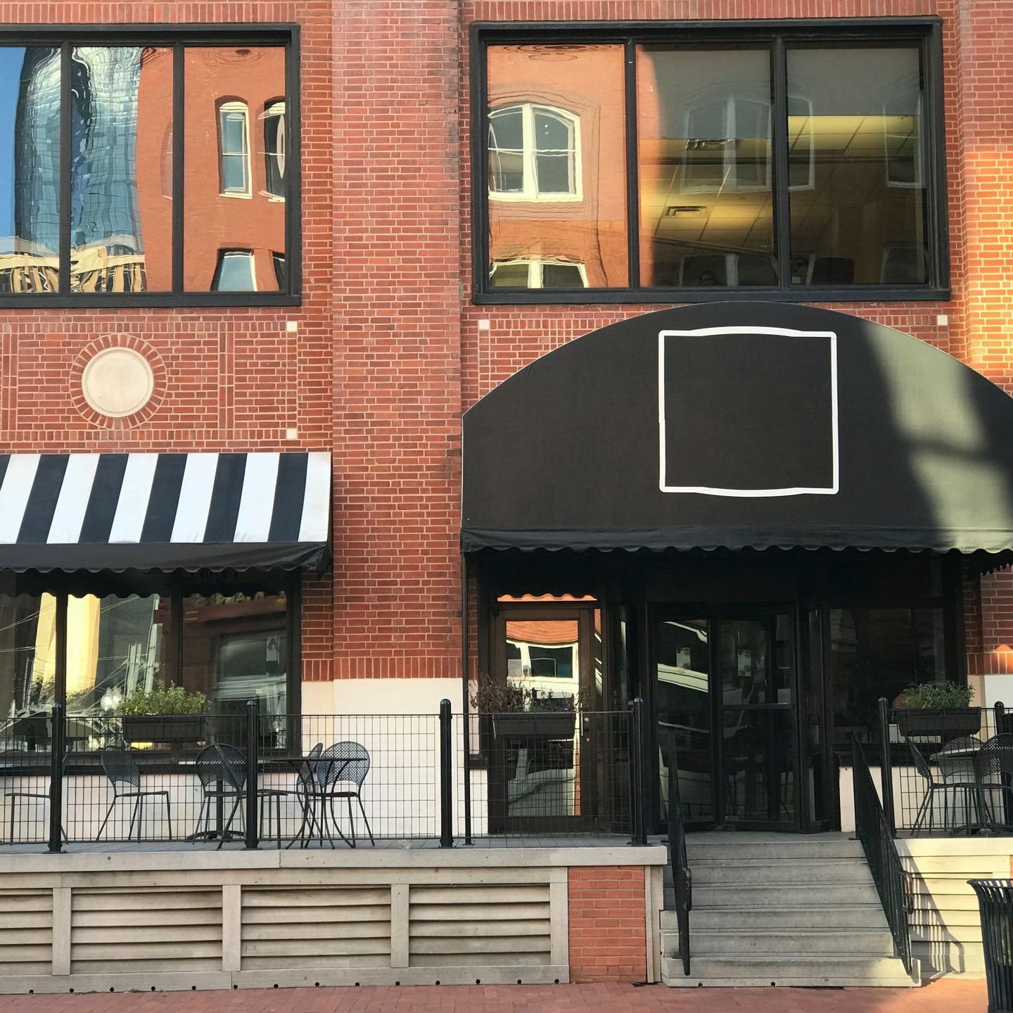Brick building with black awnings, windows reflecting other buildings, and a small outdoor seating area.