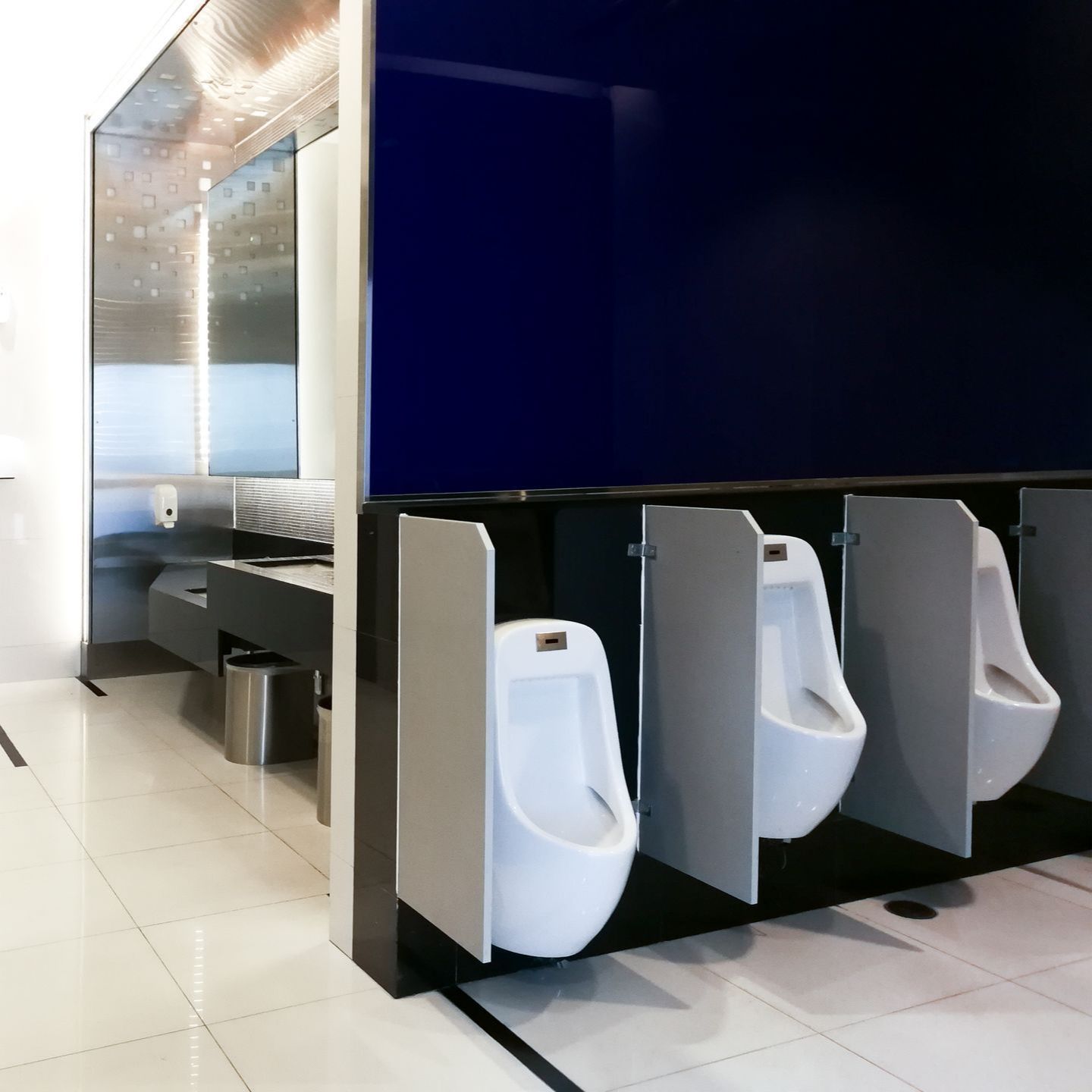 Modern men's room with three urinals and a sink area. Blue wall, white tile floor, and stainless steel accents.