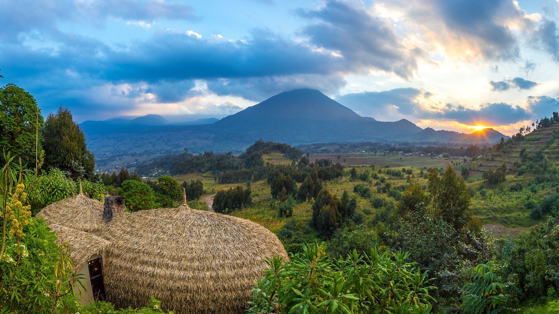 A golden sunset illuminates a mountain range above a valley with a traditional thatched-roof dwelling in the foreground.