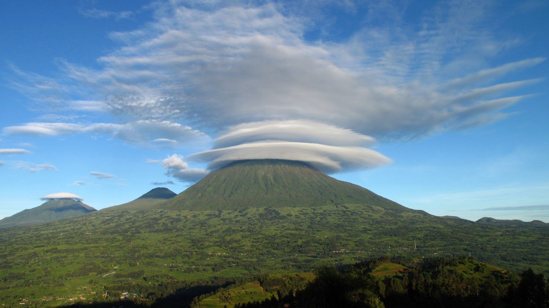 A green, cone-shaped mountain topped with layered, disc-like lenticular clouds under a bright blue sky.