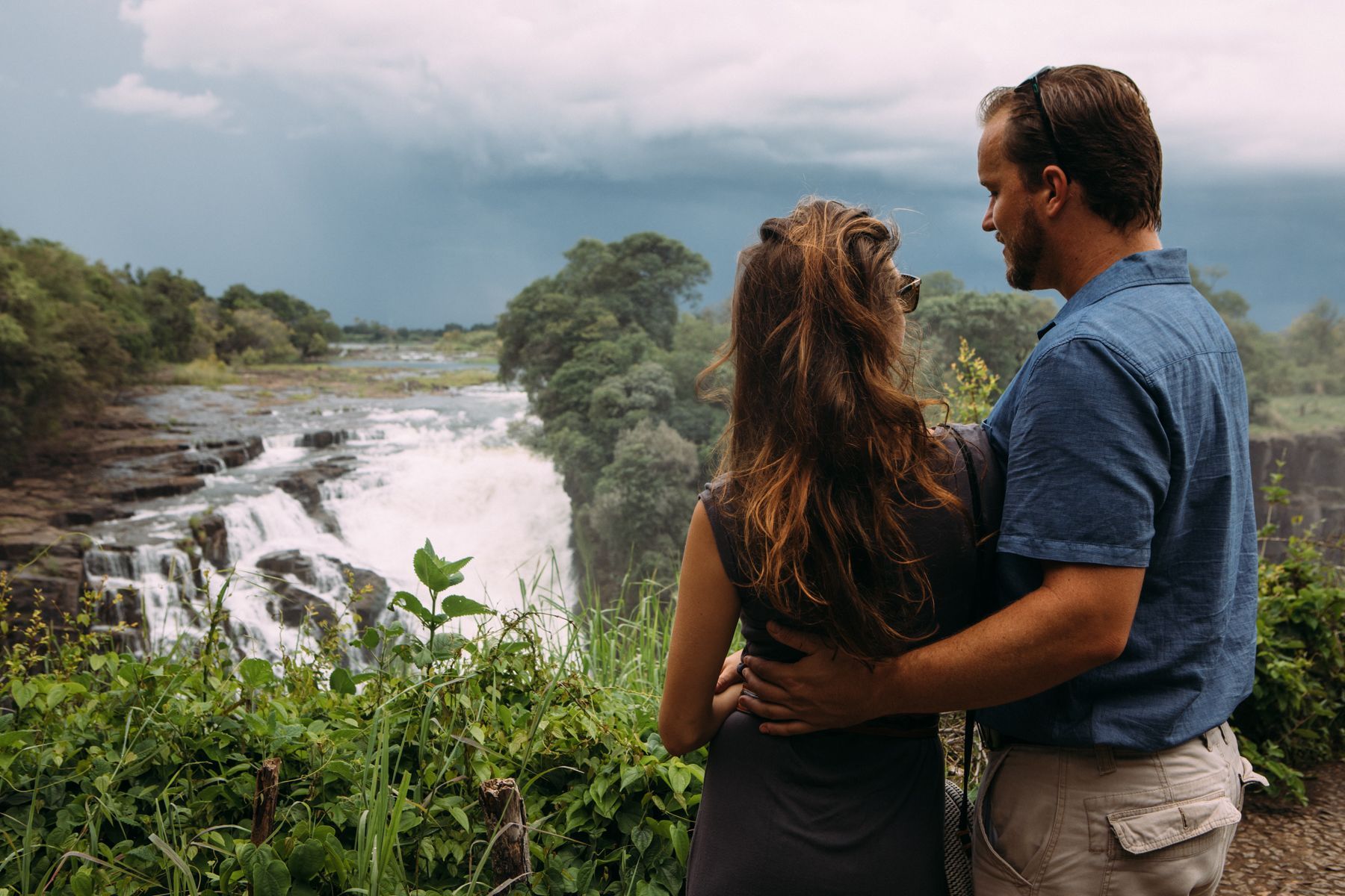 A couple stands with their arms around each other, looking at a waterfall surrounded by lush trees under a cloudy sky.