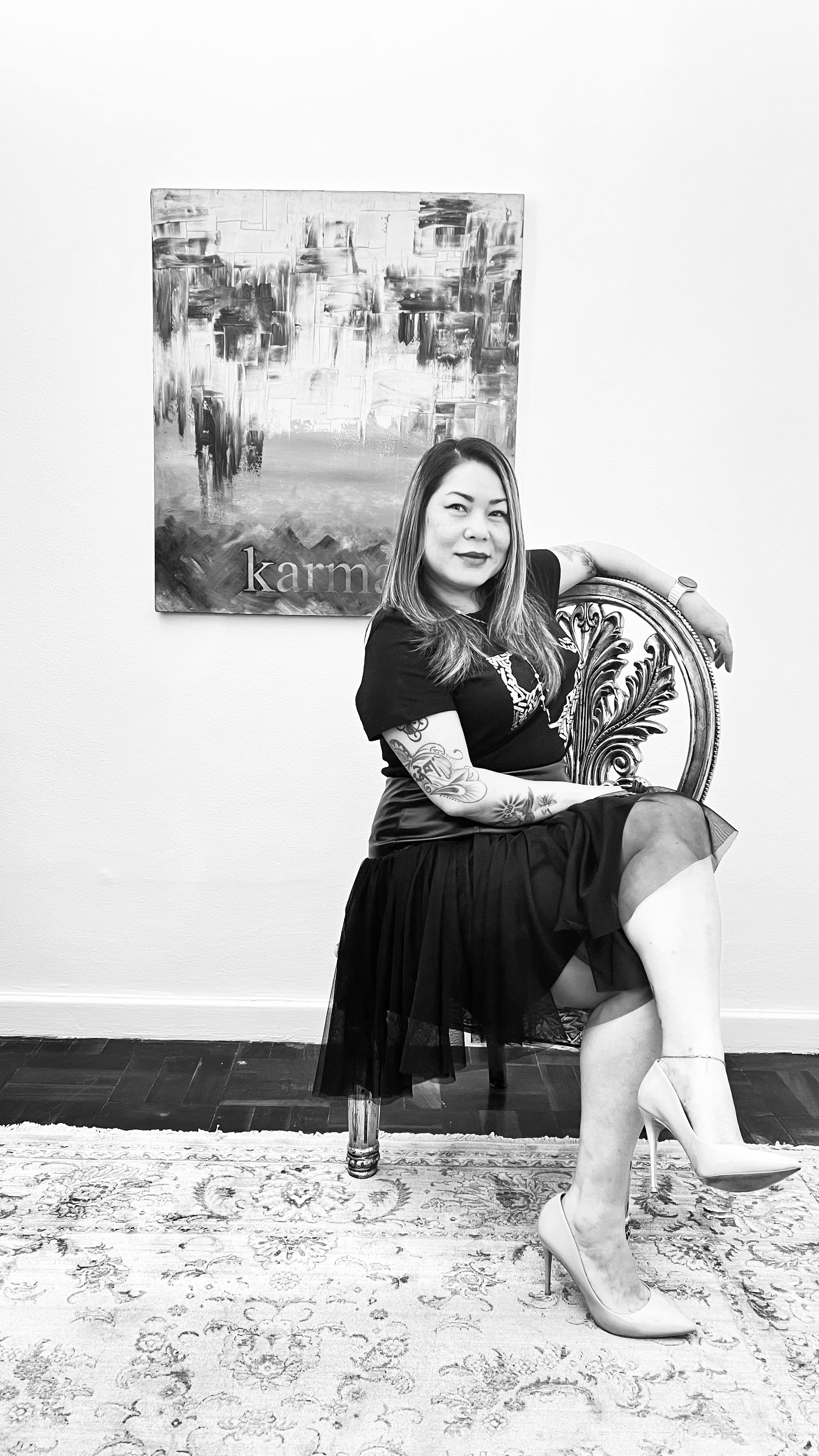 A person with tattoos sits on a chair in front of an art piece on a white wall, wearing a dark top and skirt with boots.