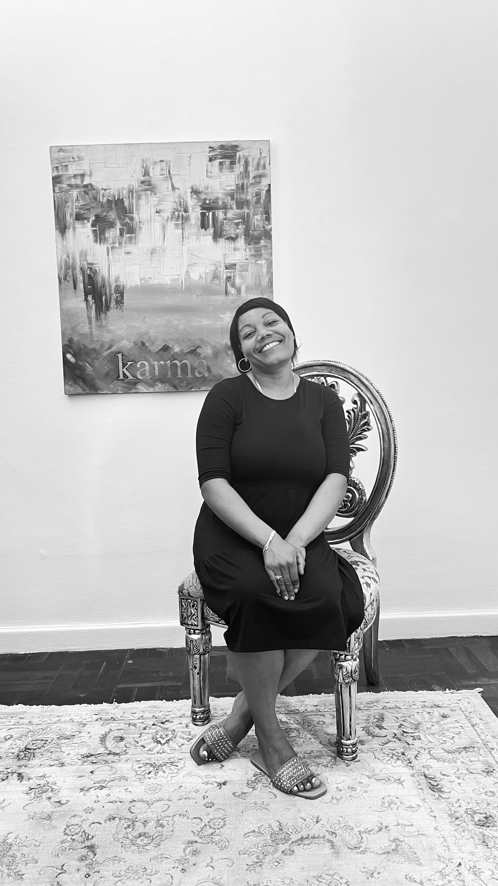 A smiling person sits on an ornate chair in front of a landscape painting, resting their feet on a patterned rug.