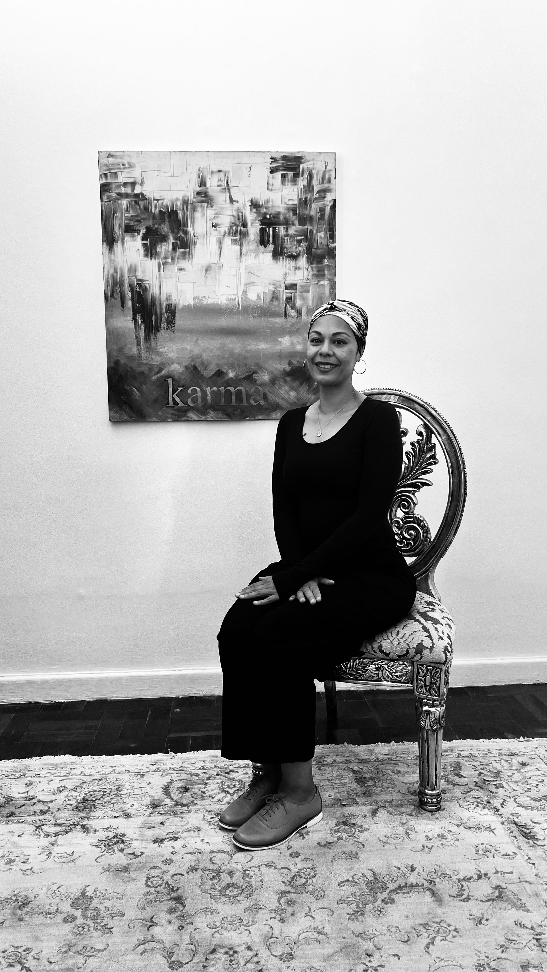 A person in black clothing sits on a patterned chair in front of a painting with the word 
