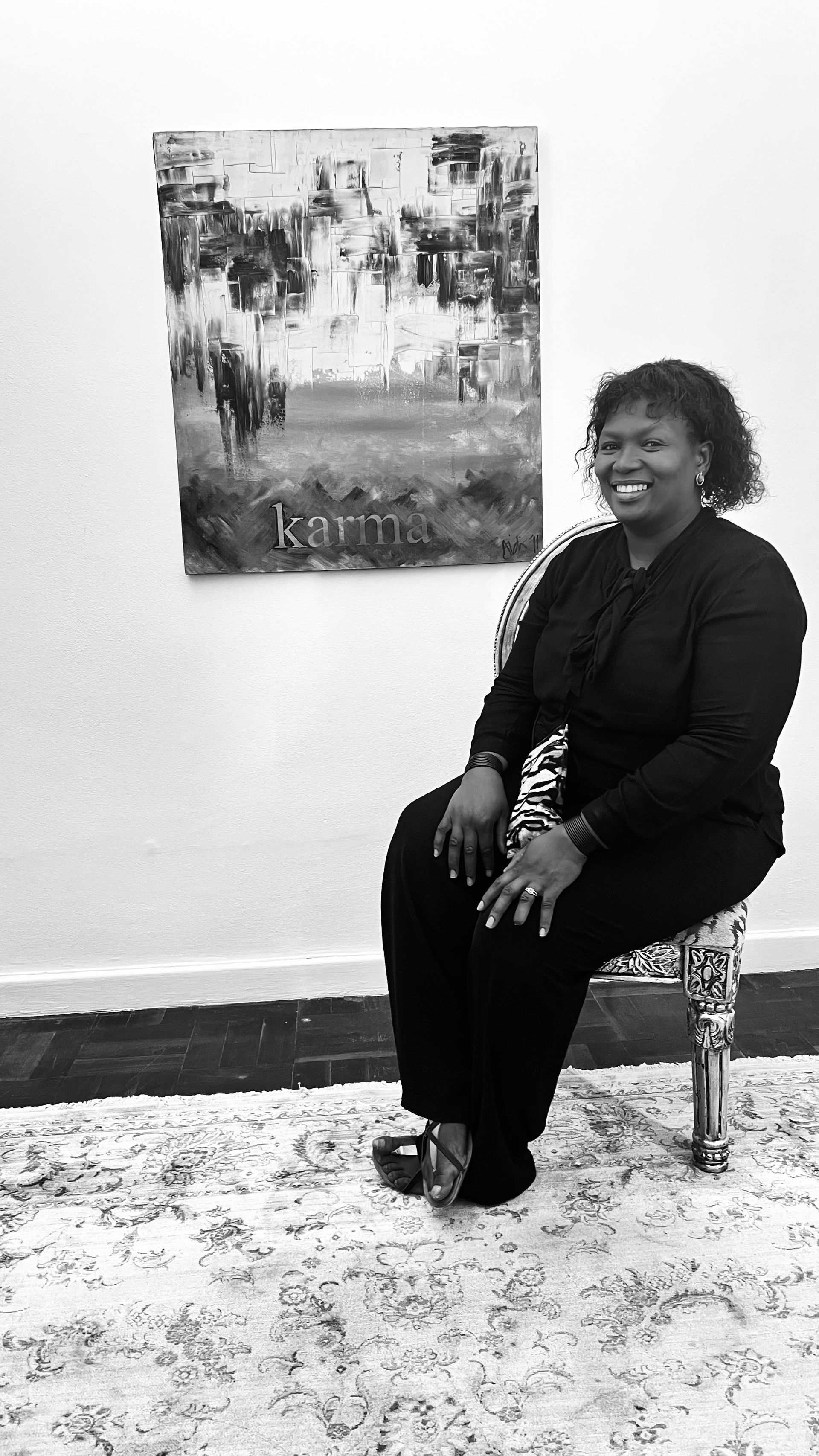 A person in black clothing sits on a patterned chair in front of a framed painting against a white wall.