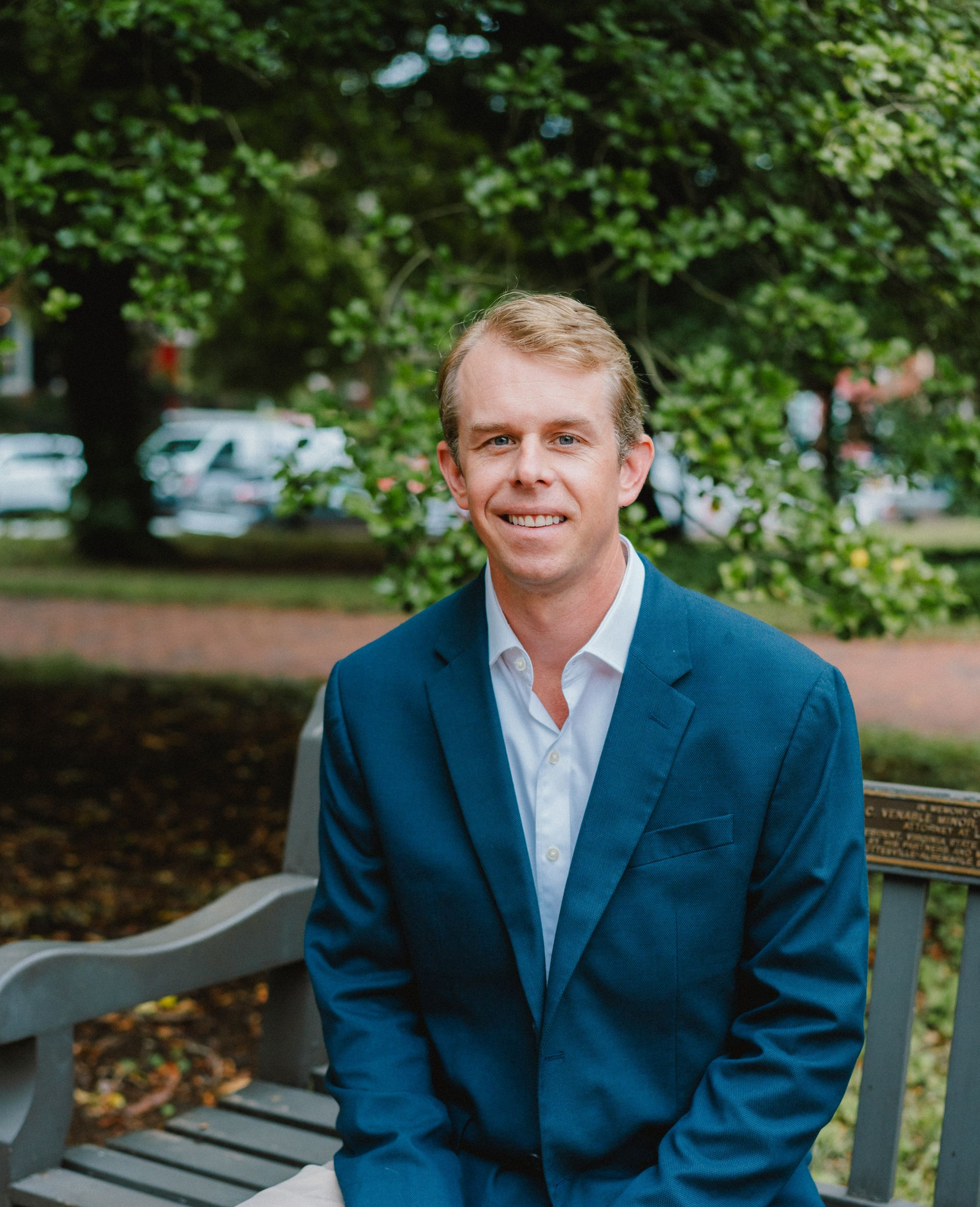 A person in a blue blazer and white shirt sitting on a wooden bench outdoors in a park-like setting.