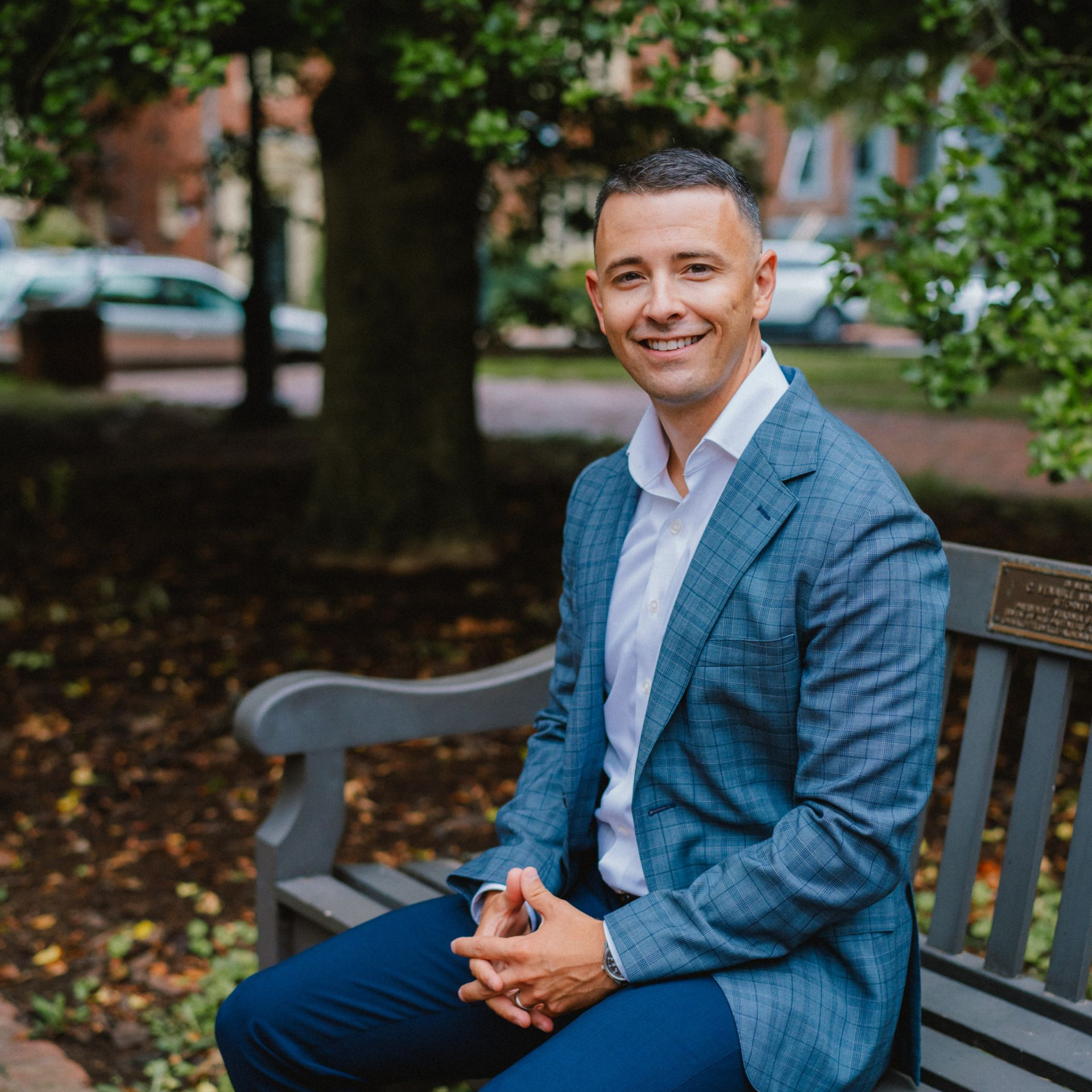 A smiling person wearing a blue plaid blazer and white shirt sits on a wooden bench outdoors in a wooded area.