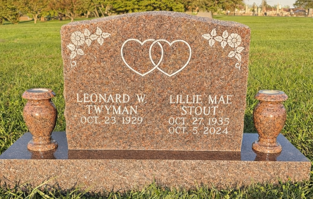A red granite headstone with two carved hearts, floral motifs, and the names Twyman and Stout, set in a grassy cemetery.