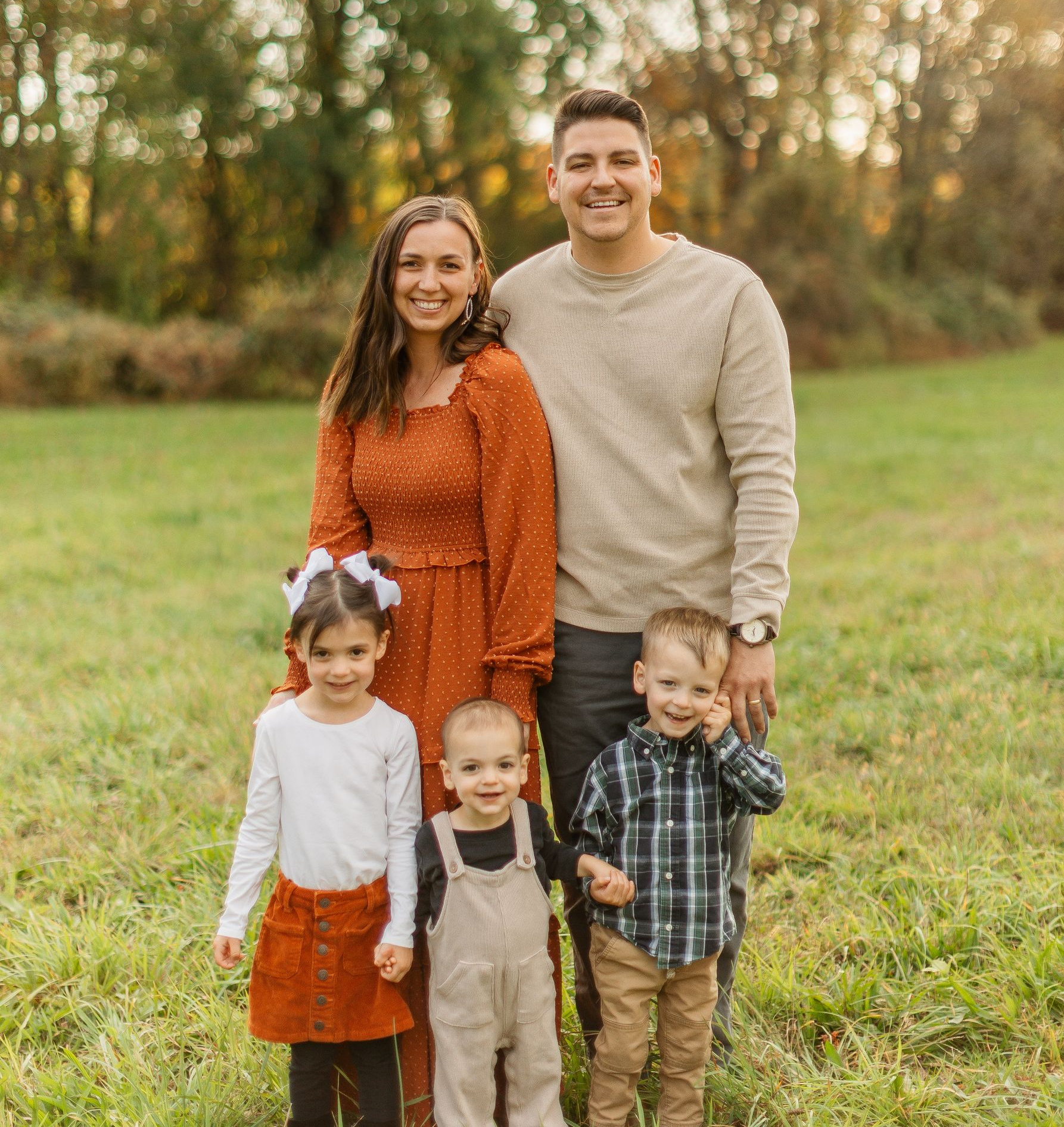 Family of five smiling outdoors: father holding a child, mother holding a baby, and a child standing in front.