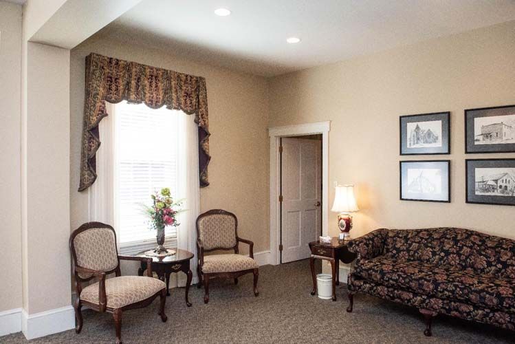 Cozy room with floral couch, two chairs, and a table. Window with floral valance, framed art, and a lamp.
