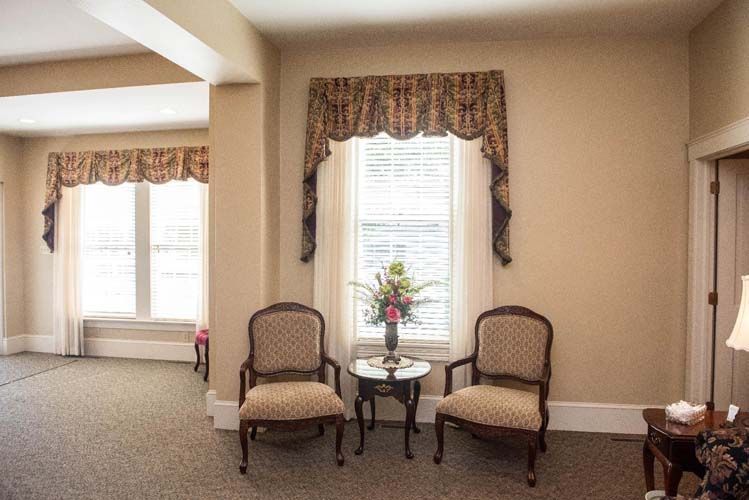 Two upholstered chairs, small table, floral arrangement, and window with ornate valance in a room.