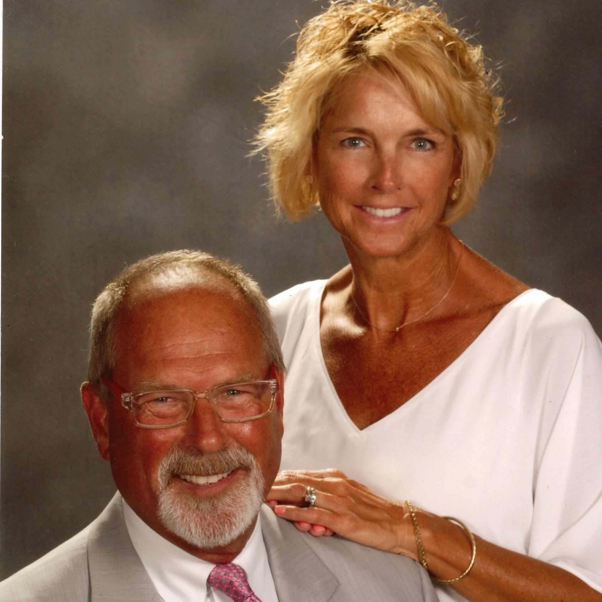 A smiling man and woman pose together. The woman's hand rests on the man's shoulder. They are in a studio setting.