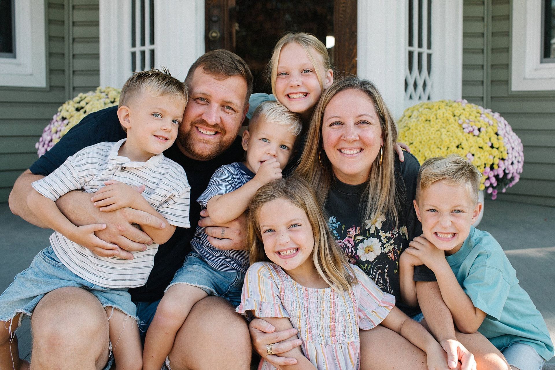 Family of eight smiling on porch steps. Two adults hold four young children; one child leans on an adult.