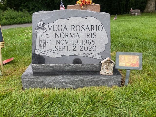 A gray granite headstone for Norma Iris Vega Rosario, featuring a lighthouse engraving, in a green grass cemetery.