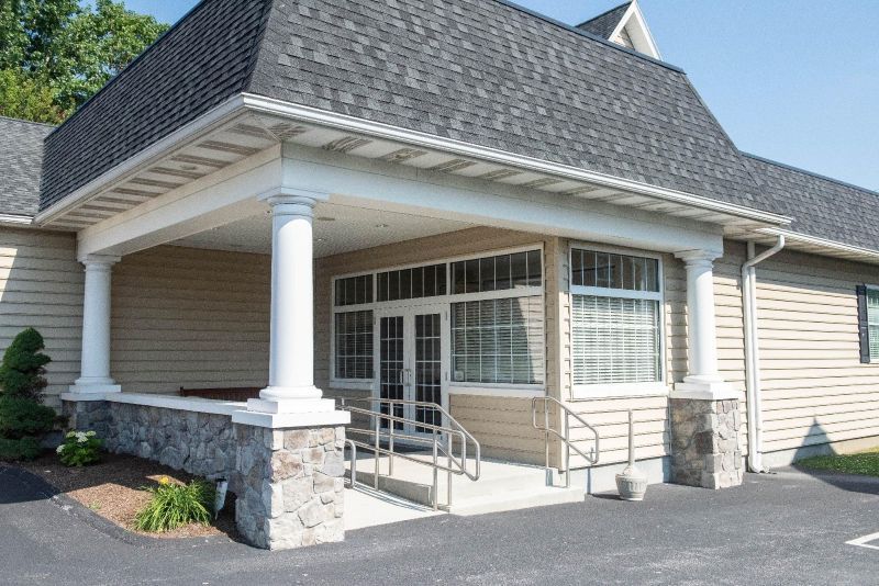 Tan building entrance with pillars, ramp, and stone facade beneath a covered porch.