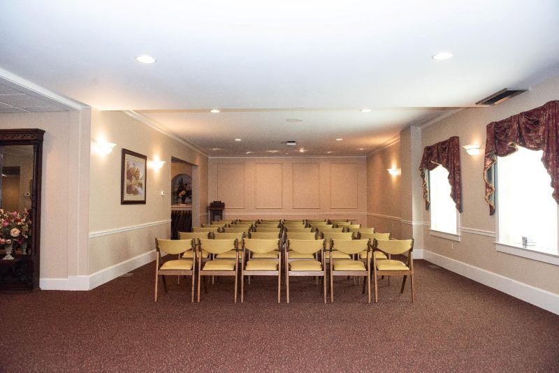 Empty beige room with rows of chairs, windows with drapes, and a dark brown carpet.