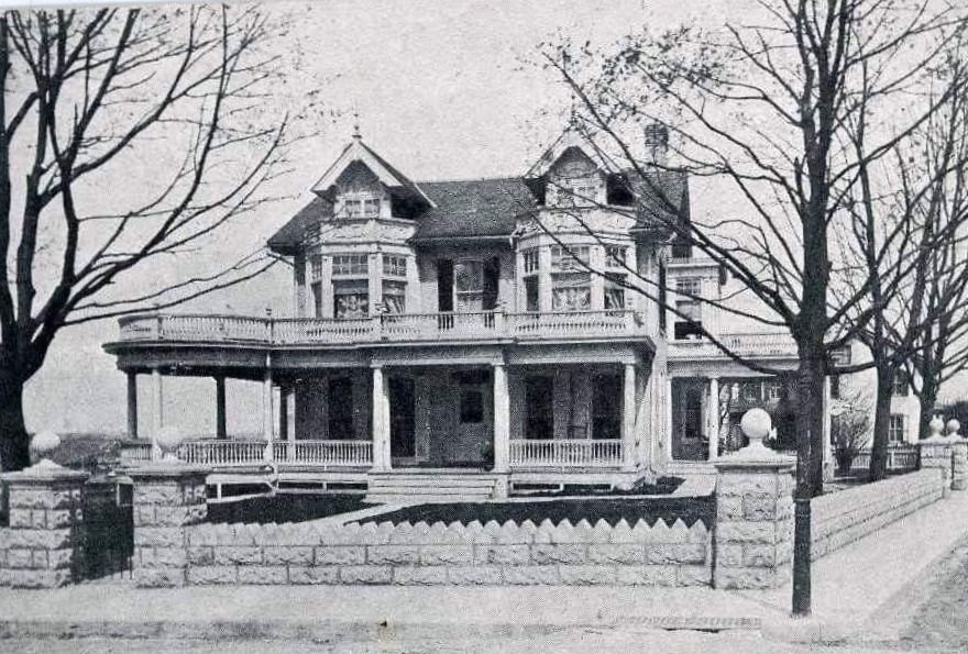Two-story Victorian house with a porch and ornate details, behind a stone wall with trees on either side.