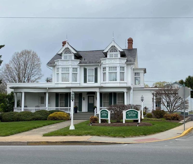 White Victorian house with green shutters, a wraparound porch, and two signs on a lawn.