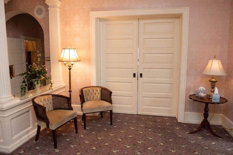 A quiet waiting area with two chairs, two lamps, and closed double doors. Soft pink walls, patterned carpet.