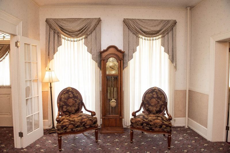 Room with ornate chairs, grandfather clock, curtains, and doorway. Beige and brown tones.