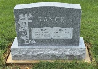 A grey granite gravestone for the Ranck family, featuring a cross and rose engraving, set in a grassy cemetery.