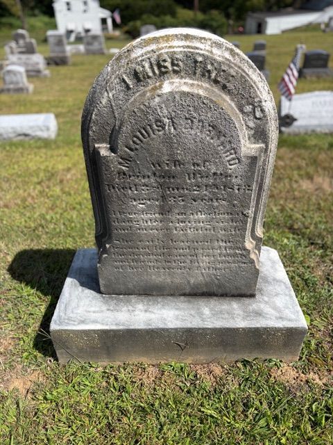 A weathered, rounded gravestone in a cemetery with a small American flag in the background.
