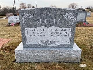 A grey granite headstone for Harold B. and Alma Mae Shultz in a cemetery, featuring an open book carving.
