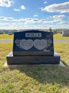 A black granite double heart headstone for the Wiker family in a sunny cemetery, inscribed with birth and death dates.